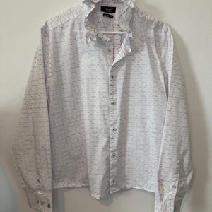 May include: White button-down shirt with a ruffled collar and long sleeves. The shirt has a subtle pattern of small blue and red dashes. The cuffs feature a small blue penguin logo. The shirt is hanging on a white hanger.