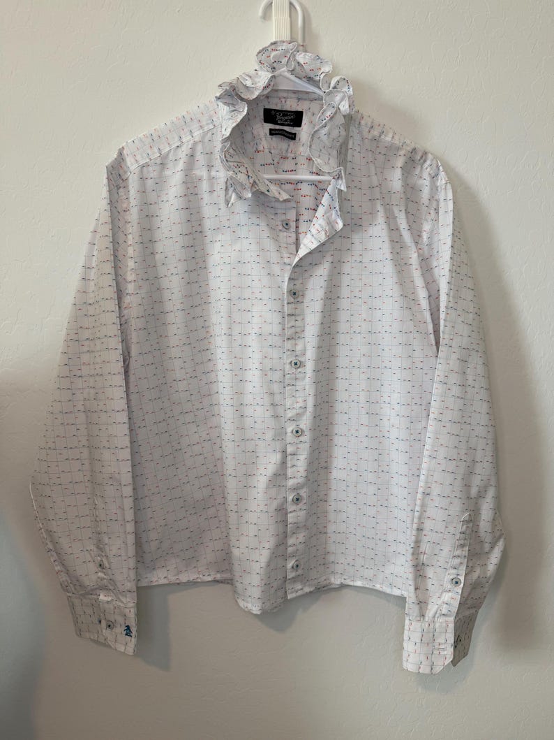 May include: White button-down shirt with a ruffled collar and long sleeves. The shirt has a subtle pattern of small blue and red dashes. The cuffs feature a small blue penguin logo. The shirt is hanging on a white hanger.