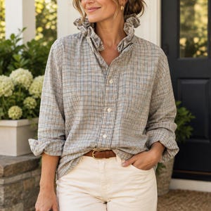 May include: A long-sleeved button-down shirt featuring a grey, tan, and blue plaid pattern with a ruffled collar. The shirt is paired with cream-coloured trousers and a brown leather belt. The sleeves are rolled up at the cuffs.
