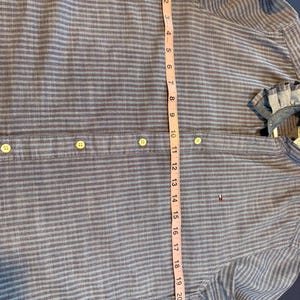 May include: Blue and gray striped button-down shirt with a measurement tape showing a length of approximately 23 inches. The shirt has a small logo on the chest and a classic collar. The shirt is made of a lightweight fabric.