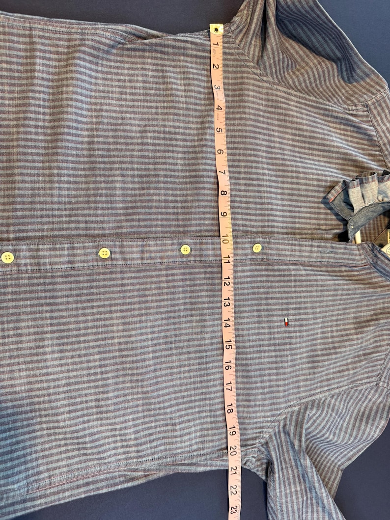 May include: Blue and gray striped button-down shirt with a measurement tape showing a length of approximately 23 inches. The shirt has a small logo on the chest and a classic collar. The shirt is made of a lightweight fabric.