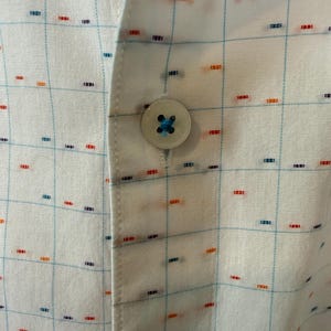 May include: Close-up of a white button-down shirt with a grid pattern. The shirt features a light blue grid design with small, colorful dashes in red, orange, and blue. A single, round button with blue thread is visible.