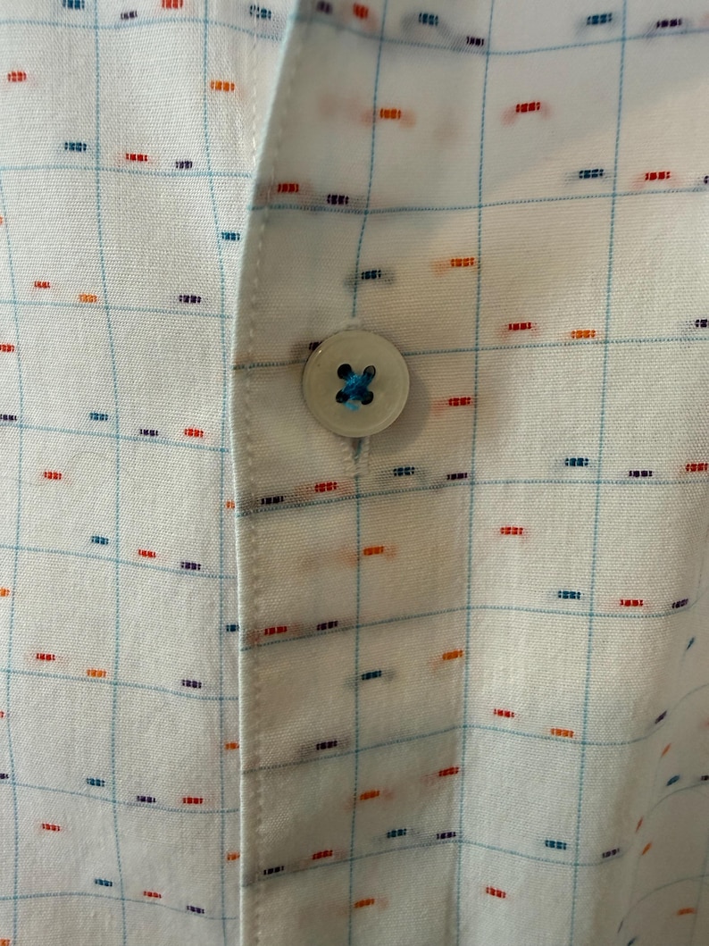 May include: Close-up of a white button-down shirt with a grid pattern. The shirt features a light blue grid design with small, colorful dashes in red, orange, and blue. A single, round button with blue thread is visible.