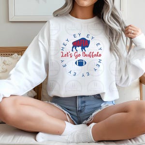 Puede incluir: Sudadera blanca con un diseño gráfico que presenta un búfalo azul, un balón de fútbol y el texto "Hey Mafia, Let's Go Buffalo" en rojo y azul. La sudadera es de manga larga.