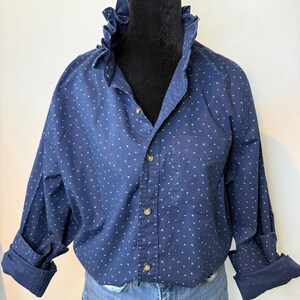 May include: A navy blue button-down shirt with a ruffled collar and small white and red triangle patterns. The long sleeves are cuffed, and the shirt has a single chest pocket. The shirt is paired with blue jeans.