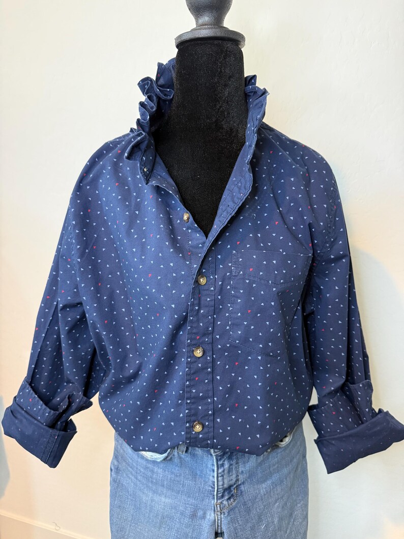 May include: A navy blue button-down shirt with a ruffled collar and small white and red triangle patterns. The long sleeves are cuffed, and the shirt has a single chest pocket. The shirt is paired with blue jeans.