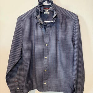 May include: A blue striped button-down shirt with a ruffled collar. The long-sleeved shirt has a vertical stripe pattern and a small logo on the chest. The shirt is hanging on a white hanger.