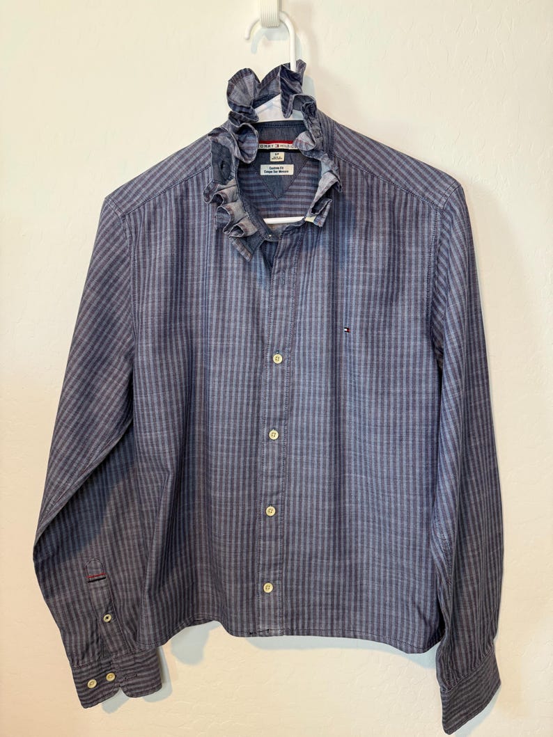 May include: A blue striped button-down shirt with a ruffled collar. The long-sleeved shirt has a vertical stripe pattern and a small logo on the chest. The shirt is hanging on a white hanger.