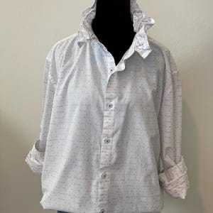 May include: White button-down shirt with a ruffled collar and cuffs. The shirt has a subtle pattern of small blue and red dots and lines. The sleeves are rolled up, revealing a patterned cuff.