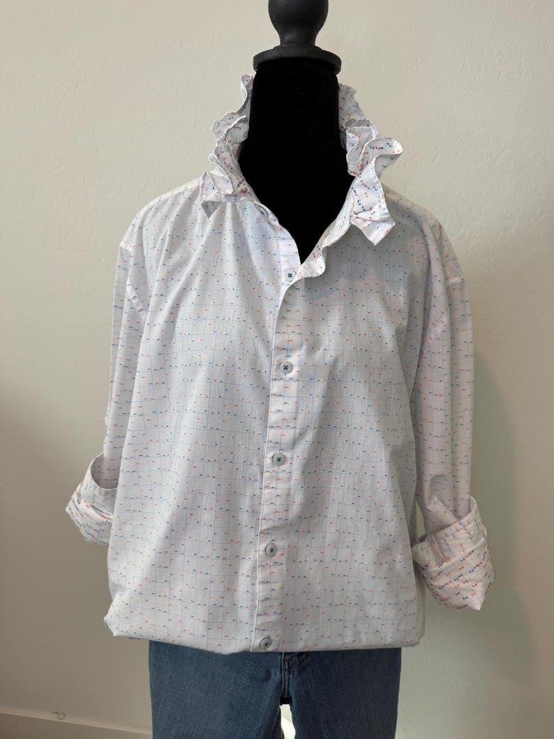 May include: White button-down shirt with a ruffled collar and cuffs. The shirt has a subtle pattern of small blue and red dots and lines. The sleeves are rolled up, revealing a patterned cuff.