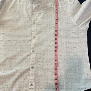 May include: White button-down shirt with a ruffled collar. The shirt has a subtle grid pattern with small red and blue accents. A pink measuring tape shows the shirt is approximately 26 inches long.