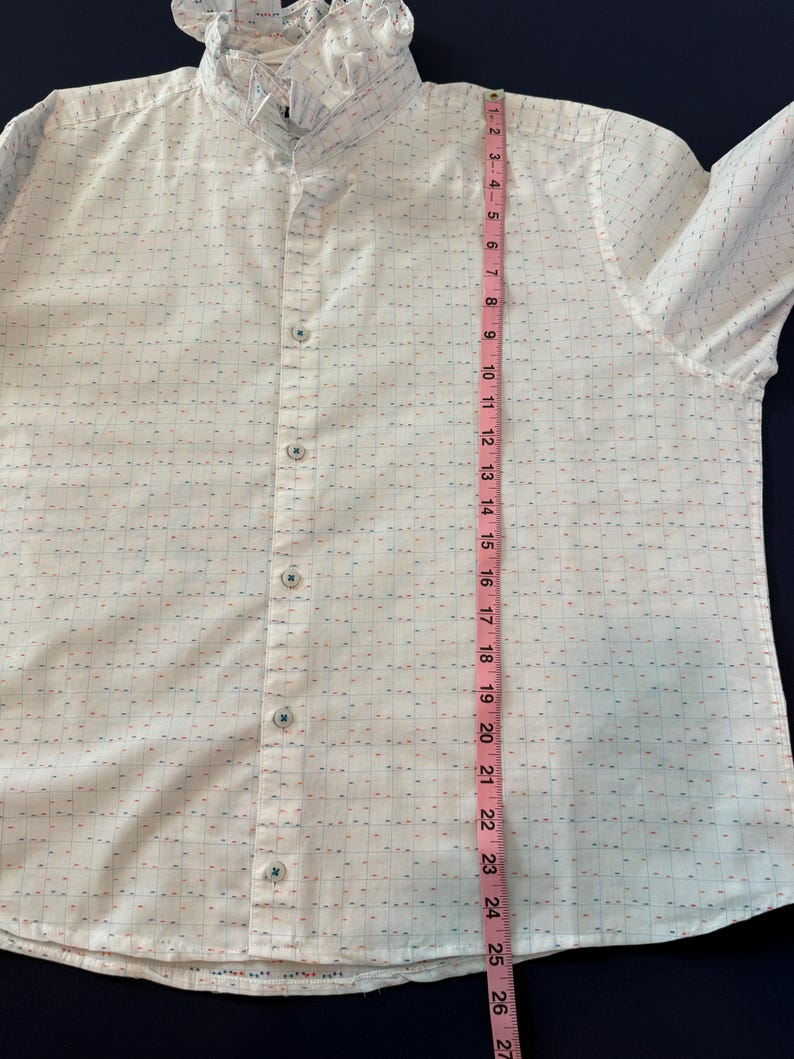 May include: White button-down shirt with a ruffled collar. The shirt has a subtle grid pattern with small red and blue accents. A pink measuring tape shows the shirt is approximately 26 inches long.