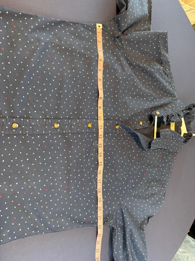 May include: A navy blue button-down shirt with a white and red triangle pattern. The shirt has gold-colored buttons and a ruffled collar. The shirt measures approximately 26 inches in length.