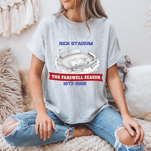 May include: A light gray t-shirt featuring a detailed black and white illustration of a stadium. The text "RICH STADIUM" is above the stadium image, with "THE FAREWELL SEASON 1973-2025" below it in red and blue.