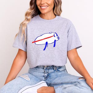 May include: A light gray t-shirt featuring a white buffalo graphic with a red stripe and blue accents. The shirt is made of a soft material and is suitable for casual wear. The graphic is a simple, cartoon-like design.