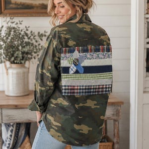 May include: A camouflage print button-down shirt featuring a large rectangular patch on the back. The patch is composed of horizontal fabric strips in various plaid, floral, and solid patterns, with a central flower applique made of fabric scraps.