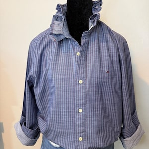 May include: A blue striped button-down shirt with a ruffled collar and rolled-up sleeves. The shirt features a small logo on the chest and is displayed on a mannequin. The shirt is paired with denim jeans.