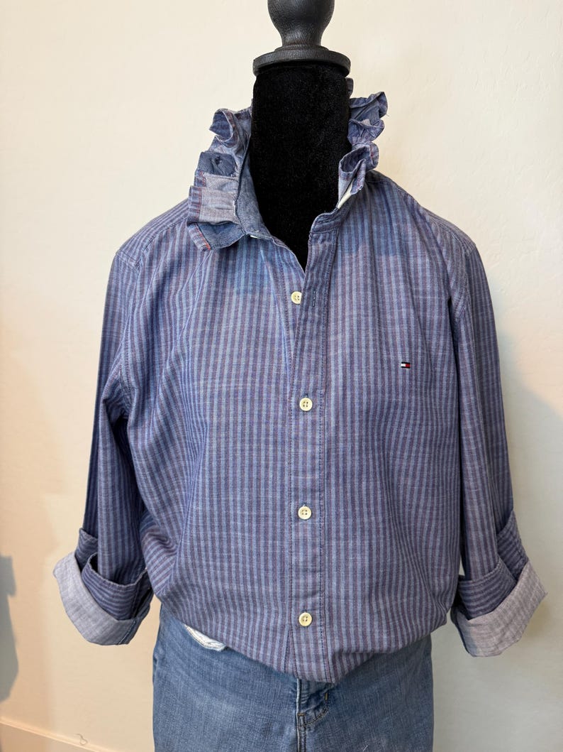 May include: A blue striped button-down shirt with a ruffled collar and rolled-up sleeves. The shirt features a small logo on the chest and is displayed on a mannequin. The shirt is paired with denim jeans.