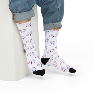 Può includere: Calzini bianchi con un motivo ripetuto di forme astratte blu e rosse. I calzini hanno talloni e dita neri. La persona indossa jeans blu.