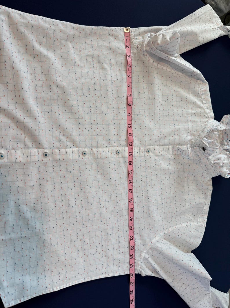 May include: White button-down shirt with a subtle grid pattern of blue, red, and pink dashes. The shirt has a ruffled collar and cuffs. A pink measuring tape shows the shirt is approximately 29 inches long.
