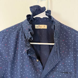 May include: Navy blue button-down shirt with a ruffled collar and a small pocket. The shirt is patterned with white and red triangles. The brand "Hollister" is visible on the label. The shirt is hanging on a white hanger.