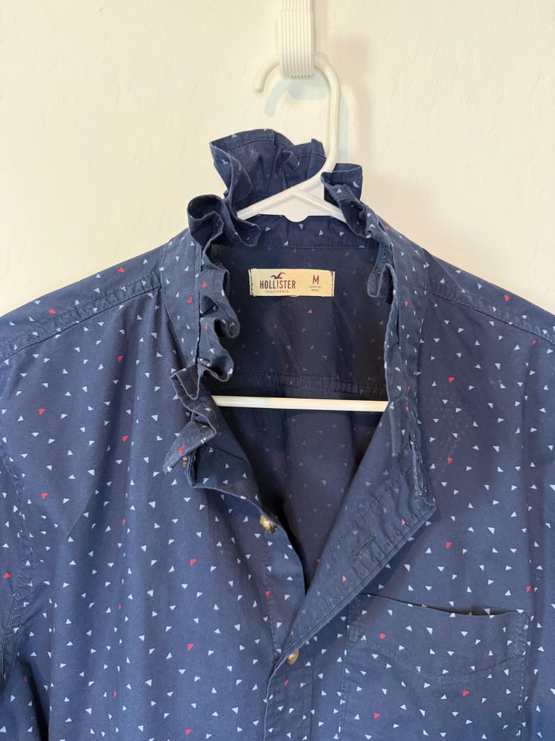 May include: Navy blue button-down shirt with a ruffled collar and a small pocket. The shirt is patterned with white and red triangles. The brand "Hollister" is visible on the label. The shirt is hanging on a white hanger.