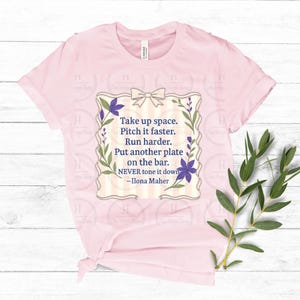 May include: A light pink t-shirt with a quote in a decorative frame. The frame has a bow at the top, floral accents, and the text: "Take up space. Pitch it faster. Run harder. Put another plate on the bar. NEVER tone it down -Ilona Maher."