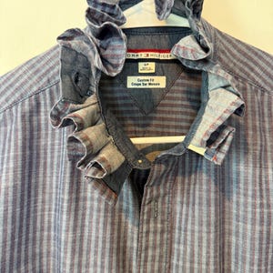 May include: A blue and red striped button-down shirt with a ruffled collar. The shirt is hanging on a white hanger. The Tommy Hilfiger logo is visible on the shirt. The shirt has a custom fit.