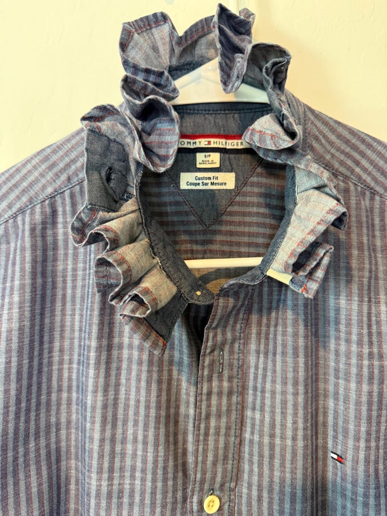 May include: A blue and red striped button-down shirt with a ruffled collar. The shirt is hanging on a white hanger. The Tommy Hilfiger logo is visible on the shirt. The shirt has a custom fit.