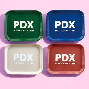 May include: Four square trays with rounded corners. Each tray is a different color: green, blue, beige, and red. The trays have the text "PDX HAVE A NICE TRIP" printed in white.