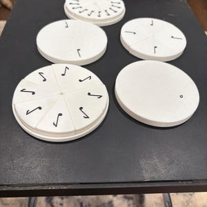 May include: A collection of white, circular objects with black markings, including musical notes and lines. Some are segmented, and others are solid. The objects are arranged on a dark surface, suggesting a game or educational tool.