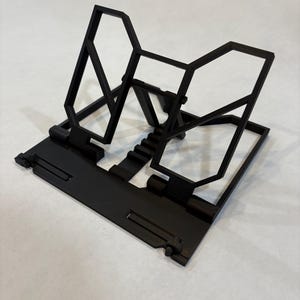 May include: A black, geometric phone stand. The stand features a rectangular base with a series of angled supports and a central platform. The design incorporates hexagonal and rectangular shapes, creating a modern aesthetic. The stand is made of a solid, matte black material.