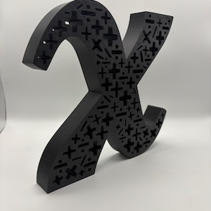 May include: A large, black, three-dimensional letter "X" with a matte finish. The letter is filled with cut-out mathematical symbols, including plus signs, minus signs, and division symbols. The "X" is standing upright against a white background.