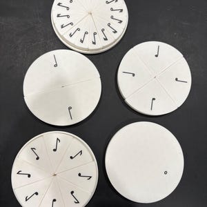May include: A collection of white, circular music-themed educational toys. Some have black musical notes around the edges, while others are divided into segments with single notes. The toys are arranged on a dark surface.