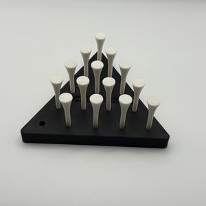 May include: A black triangular golf tee holder with 15 white golf tees. The holder is made of a solid material and has a small hole on one side. The tees are arranged in a pyramid shape.