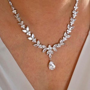 May include: A silver-toned necklace with a floral design. The necklace features clear, sparkling stones set in a leaf-like pattern, with a teardrop-shaped pendant. The necklace is worn around the neck.