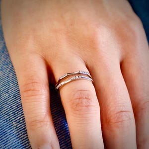 May include: A silver ring with a double band design, adorned with small, sparkling stones. The ring is worn on a finger, showcasing its delicate and elegant style. The ring's design is a wave-like pattern.