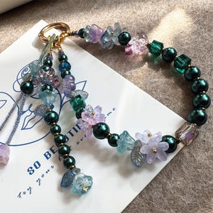 Puede incluir: Un llavero de cuentas con un anillo dorado, con una mezcla de cuentas verdes, azules y moradas. El diseño incluye detalles de flores y hojas, con una borla colgante. El texto "SO BEAUTIFUL" es visible.