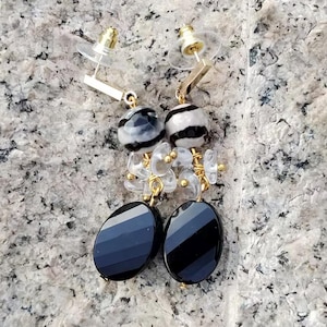 May include: A pair of dangle earrings featuring black and white striped beads, clear crystal accents, and oval black gemstones. The earrings have gold-colored metal accents and post-style closures.