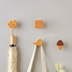 Può includere: Un set di quattro ganci da parete in legno a forma di cibo, tra cui un biscotto con gocce di cioccolato, un cracker quadrato, una fetta di formaggio svizzero e un cono gelato al cioccolato, montati su una parete con un ombrello e una borsa.