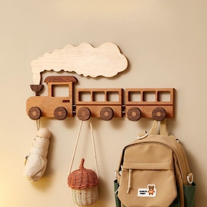 Puede incluir: Perchero de pared con forma de tren de madera con un acento de nube. El tren marrón tiene tres vagones con ruedas circulares y ganchos para colgar objetos. Un paraguas, una cesta tejida y una mochila cuelgan de él.