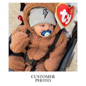 May include: A baby wearing a brown teddy bear-style hooded fleece jumpsuit and a gray beanie with a black lightning bolt graphic. The baby has a blue and white pacifier and a red heart-shaped Ty tag attached to the hood of the jumpsuit.