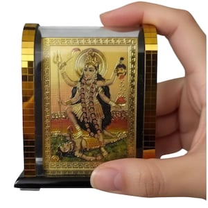 May include: A gold-colored religious icon featuring a multi-armed deity with a trident, set within a clear acrylic block. The block has gold-colored mosaic tile accents on the sides and a black base. The icon is held in a hand.