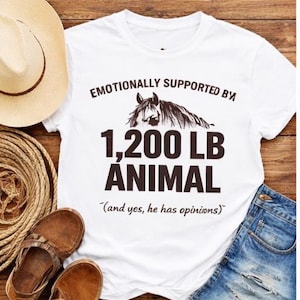 Może przedstawiać: Biały ceramiczny kubek z czarnym uchwytem i napisem "But First, Coffee" pogrubioną czcionką. Kubek stoi na drewnianej powierzchni. Kubek ma około 10 cm wysokości.