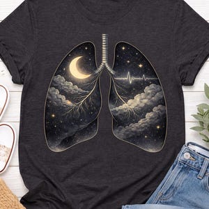 Könnte beinhalten: Ein dunkelgraues T-Shirt mit einem grafischen Aufdruck menschlicher Lungen. Das Innere der Lungen zeigt eine nächtliche Himmelsszene mit einer Mondsichel, Sternen, flauschigen Wolken und einer weißen EKG-Herzschlaglinie auf der rechten Seite.