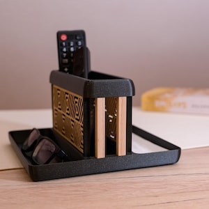 Könnte beinhalten: Ein schwarzer und goldfarbener Schreibtisch-Organizer aus Holz mit einem Tablett für Brillen und einem Fach für eine Fernbedienung. Der Organizer hat ein geometrisches Muster an der Seite und zwei Holzteilern.