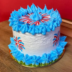 May include: A small round cake with white frosting, decorated with bright blue piped icing swirls along the top and bottom edges. The sides and top feature red and blue firework-style patterns, with a single blue icing dollop in the center.
