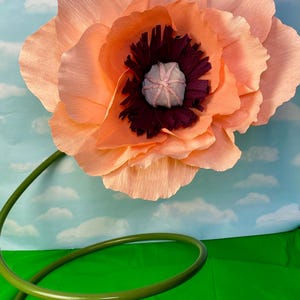 May include: A large, peach-colored paper flower with a dark burgundy center and a light pink center. The flower has a green stem that curves into a spiral. The background is a blue sky with white clouds and a green surface.