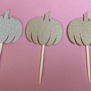 May include: Three glittery pumpkin-shaped cupcake toppers. Each pumpkin is a different shade of gold and silver, with a wooden stick attached. The toppers are set against a pink background, perfect for fall-themed events.