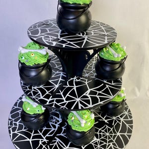 May include: A three-tiered black and white spiderweb-themed cupcake stand. Each tier holds black cauldron-shaped containers filled with green frosted cupcakes, decorated with bone-shaped candies and candy eyeballs. A festive Halloween dessert display.